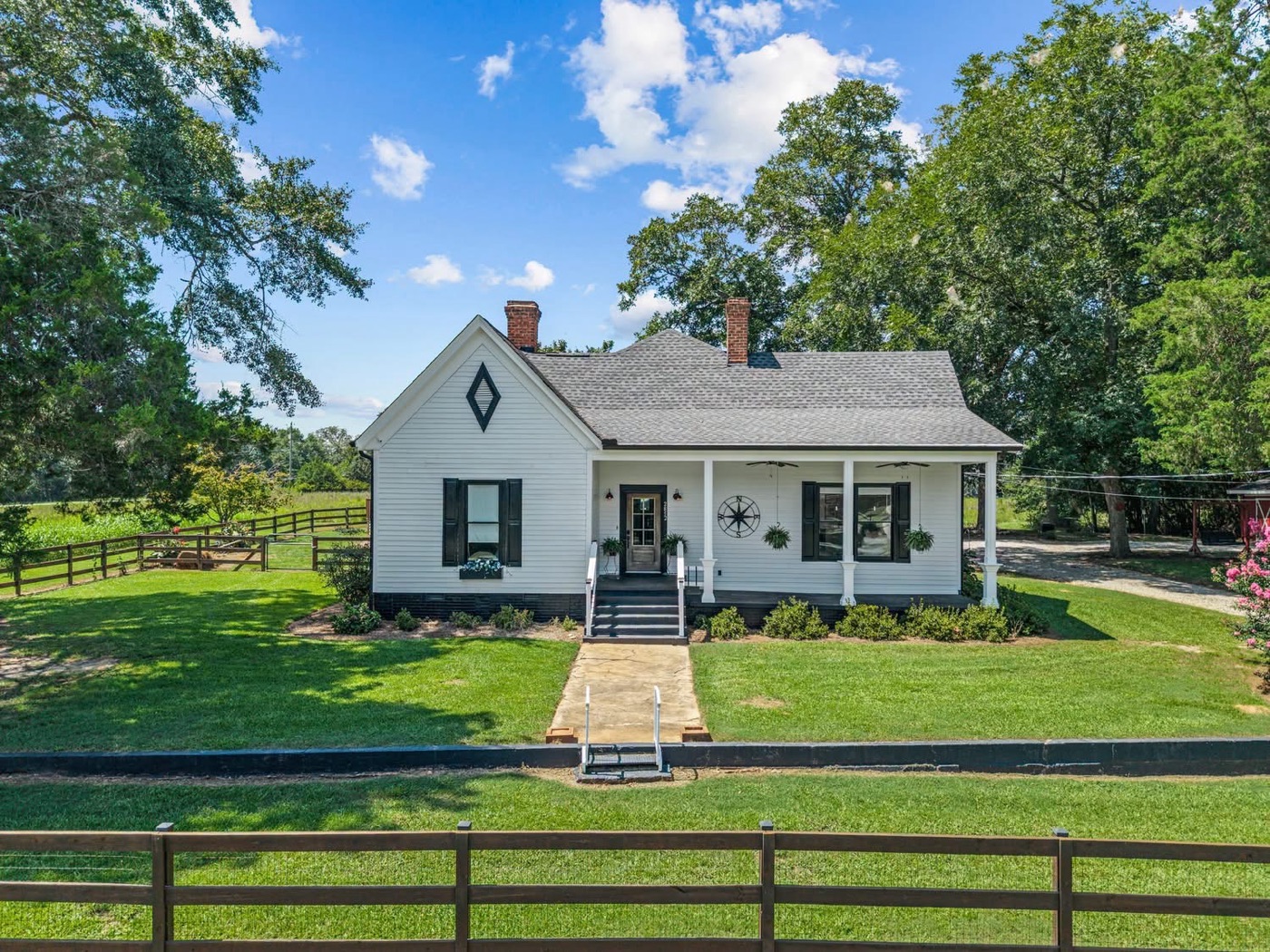 A white farmhouse with black shutters and a front porch, behind a board fence and a green lawn