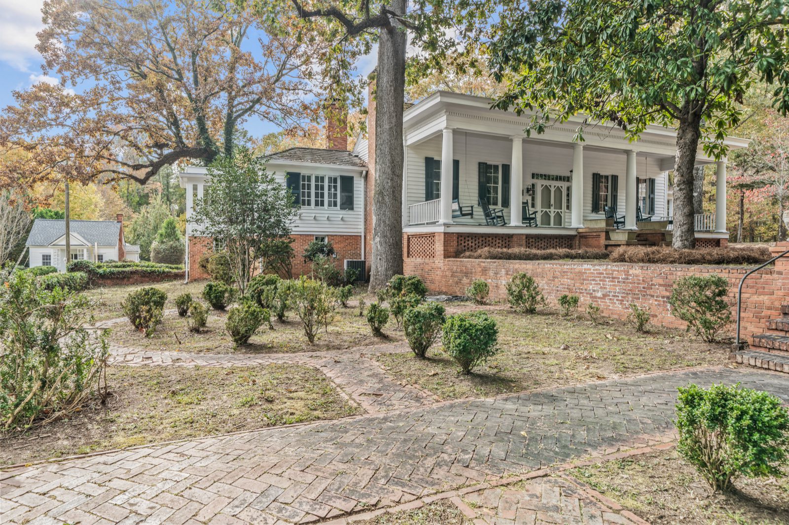 A white historic house with a columned porch above a brick wall, a herringbone brick walk and boxwoods under autumn oaks