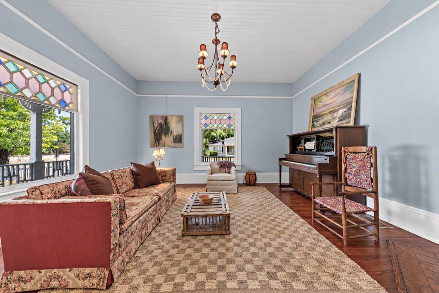 A sunny front parlor with pale blue walls, stained-glass window transoms and an upright piano